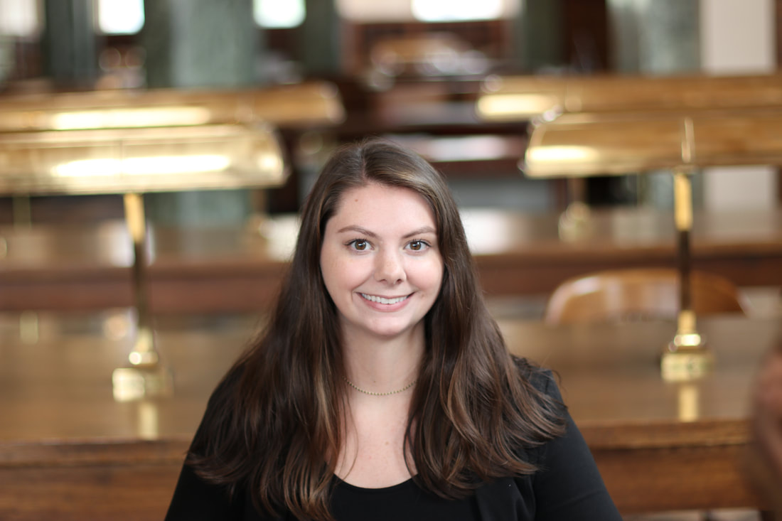 Photo of Savannah Foreman. She is sitting in a library. She has dark hair, dark eyes, and is smiling. She is dressed in a black shirt with a black blazer and wears a thin gold necklace.